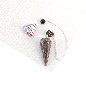May include: A dowsing pendulum featuring a faceted, speckled dark purple and grey stone point topped with a small black cat-shaped bead. A silver-toned chain connects the stone to a triangular, translucent light blue glass bead with red swirls.