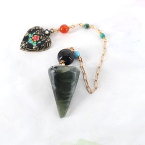 May include: A pendulum featuring a dark green, cone-shaped stone suspended from a gold-toned chain. The chain includes a black bead, small turquoise beads, an orange bead, and a heart-shaped charm with a black background and floral design.