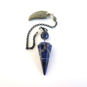May include: A pendulum with a dark blue, faceted crystal point. The pendulum has a dark chain, a blue sphere, and a decorative wing-shaped element. The crystal point is approximately 2 inches long.