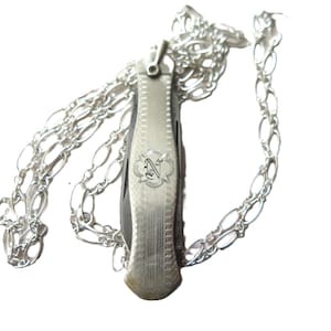 May include: A silver pocket knife pendant with a decorative engraved design and a silver chain. The knife has a detailed pattern and an emblem with the letter "N". The chain is made of oval links.