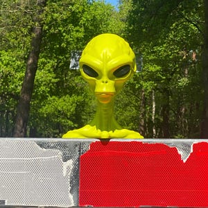 Könnte beinhalten: Eine leuchtend gelbe Alien-Kopfskulptur mit schwarzen Augen und einem breiten Mund, platziert auf einer rot-weißen reflektierenden Straßenbarriere.