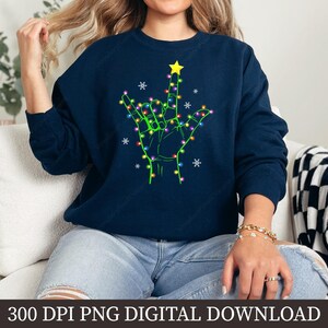 May include: Navy blue sweatshirt featuring a hand gesture with a star on top, outlined with green and decorated with colorful lights and snowflakes. The text "300 DPI PNG DIGITAL DOWNLOAD" is at the bottom. A cozy and festive design.