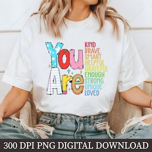 May include: A white t-shirt with a colorful graphic that says "You Are" with a list of positive words including kind, brave, smart, helpful, grateful, enough, strong, unique, and loved.