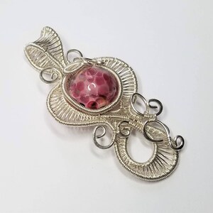 May include: A silver wire wrapped pendant with a pink and white speckled stone in the center.