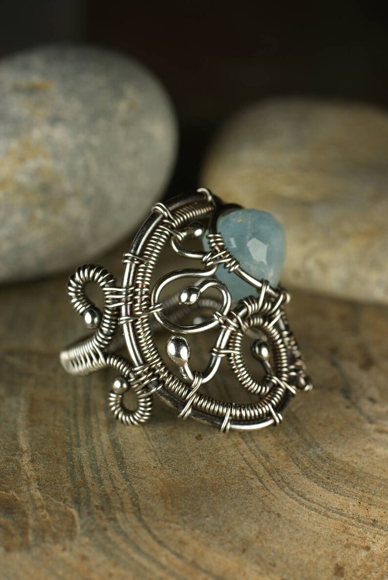 May include: A silver wire wrapped ring with a light blue gemstone. The ring is intricate and detailed, with a delicate design.