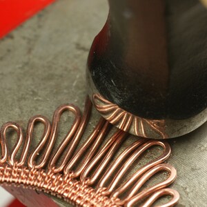 May include: A close-up of a copper wire jewelry piece being hammered with a metal mallet. The jewelry piece is made of many small loops of copper wire, creating a textured and intricate design.
