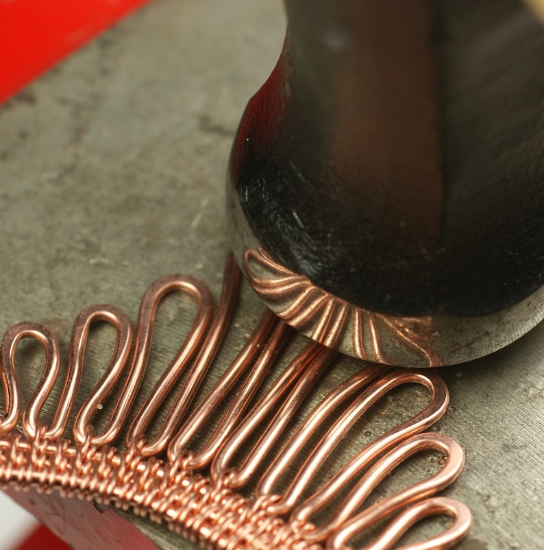 May include: A close-up of a copper wire jewelry piece being hammered with a metal mallet. The jewelry piece is made of many small loops of copper wire, creating a textured and intricate design.