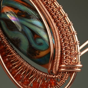 May include: A copper wire wrapped pendant featuring a colorful glass bead and orange carnelian beads. The pendant is made with a woven wire technique.