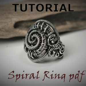 Puede incluir: Un anillo de alambre plateado con un diseño en espiral. El anillo está sobre una superficie de madera. El texto "Spiral Ring pdf" está escrito debajo del anillo.