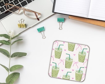 May include: Square coaster featuring a pattern of green matcha lattes with pink bows and hearts on a white background. The coaster is on a white desk with office supplies and a laptop.