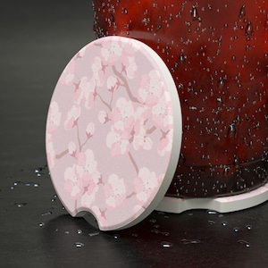 May include: A round car coaster with a floral cherry blossom design in pink and white. The coaster has a light pink background and a white border. The coaster is next to a red glass with water droplets.