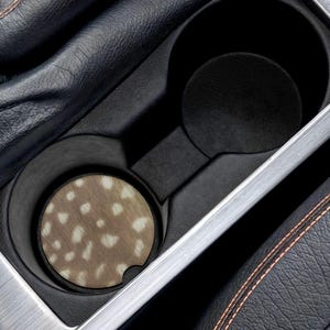 Könnte beinhalten: Ein Getränkehalter im Auto mit einem Untersetzer mit Hirschfellmuster. Der Untersetzer ist rund mit einem braunen und weißen Fleckenmuster. Der Getränkehalter ist schwarz und befindet sich in einer Autokonsole.