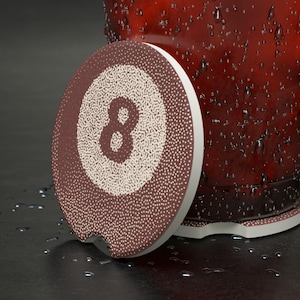 May include: A round car coaster with a brown and white 8-ball design. The coaster has a white border and a small notch for easy removal. The coaster is next to a red glass with condensation.