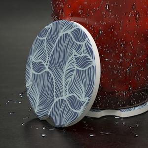May include: A set of two car coasters with a blue and white abstract leaf design. The coasters are round with a small notch and a white border. The coasters are next to a red cup with water droplets.