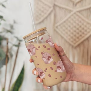 Puede incluir: Un vaso de vidrio con tapa marrón y una pajita. El vaso está lleno de un líquido marrón y tiene un patrón de cajas de leche rosas con leche de chocolate dentro.