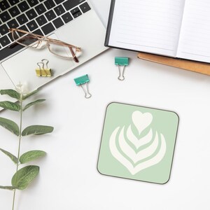 May include: A square, pale green coaster with a white latte art design of a heart and leaves. The coaster sits on a white surface with a laptop, glasses, binder clips, and a sprig of greenery.