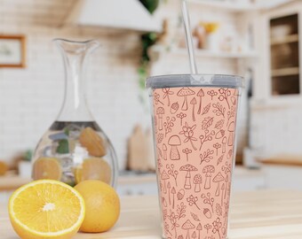 Op de afbeelding: Een roze herbruikbare beker met een rietje en een paddenstoel- en bladmotief. De beker staat op een houten oppervlak met sinaasappels op de achtergrond.