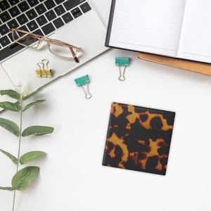 May include: A square coaster with a tortoiseshell pattern in shades of brown and black. The coaster is on a white surface, along with a laptop, glasses, binder clips, and a notebook.