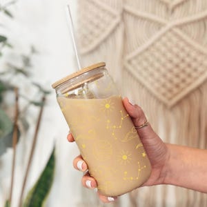 May include: A glass tumbler with a straw and a wooden lid. The tumbler is filled with a light brown liquid and has a design of yellow stars, planets, and clouds on the side.
