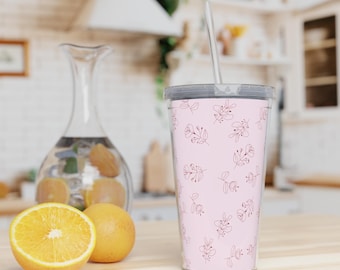 Op de afbeelding: Een roze beker met een bloemenpatroon en een doorzichtig rietje. De beker staat op een houten oppervlak met een doorgesneden sinaasappel en een hele sinaasappel. Een glazen karaf met sinaasappels staat op de achtergrond.