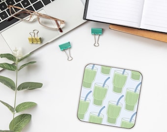 May include: Square coaster featuring a light blue backdrop and a repeating pattern of green smoothie glasses with blue straws. The coaster is on a white desk with a laptop, glasses, and stationery.