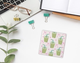 Puede incluir: Posavasos cuadrado con fondo rosa claro con un patrón de bebidas heladas verdes con pajitas, lazos rosas y pequeños corazones rosas. El posavasos está sobre una superficie blanca con un portátil, gafas y clips de encuadernación.