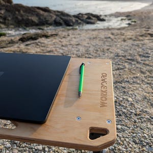 May include: A wooden workboard with a black laptop and a green pen on a pebble beach. The workboard has the word "WORKBOARD" engraved on it. The workboard has black legs and is secured with silver screws.