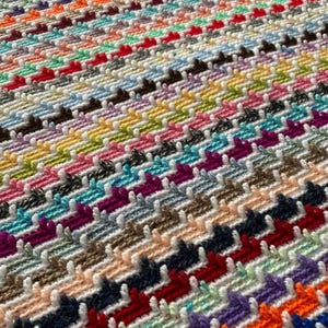 May include: A colorful crocheted blanket with a chevron pattern. The blanket is made with a variety of different colored yarns, including red, orange, yellow, green, blue, purple, and white.