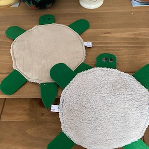 Puede incluir: Dos posavasos hechos a mano con forma de tortuga. Cada posavasos presenta un centro redondo, beige o gris, con apéndices verdes en forma de aleta. Las tortugas tienen ojos pequeños, redondos y negros. Los posavasos están sobre una superficie de madera.