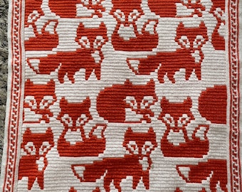 May include: A handmade crochet blanket featuring a repeating pattern of red foxes on a white background. The blanket has a red and white border. The foxes are in various poses, creating a playful and cozy design. This blanket is perfect for a nursery or as a decorative throw.