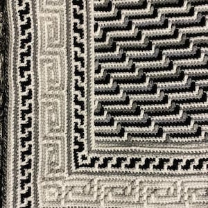 May include: A close-up of a crocheted blanket with a geometric pattern in shades of gray and black. The blanket has a border with a repeating design.