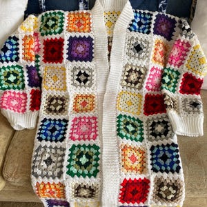 Può includere: Un cardigan bianco all'uncinetto con un davanti lungo e aperto e toppe colorate a quadrati della nonna. Il cardigan ha maniche lunghe e una vestibilità rilassata.