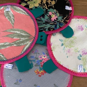 Puede incluir: Una colección de artículos de tela redondos con estampados florales y de hojas. Cada artículo tiene un borde rosa brillante y detalles en verde. Las telas presentan varios diseños, incluyendo hojas, flores y un estampado floral negro.