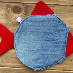 Op de afbeelding: Een handgemaakt visvormig item met een denim lijf en rode viltvinnen en kop. De vis heeft een zwart oog en een klein label. Waarschijnlijk een decoratief stuk of speelgoed.