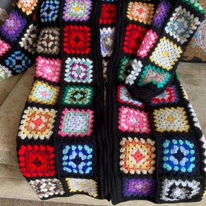 Puede incluir: Una larga chaqueta de punto de ganchillo negra con un patrón de cuadrados de la abuela de colores. La chaqueta tiene cuello en V y mangas largas.