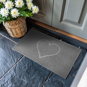 Puede incluir: Una alfombra de entrada gris con un diseño de corazón blanco. La alfombra está hecha de un material texturizado y se coloca sobre un suelo de baldosas grises.
