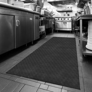 May include: Black rectangular anti-fatigue mat with a diamond pattern in a commercial kitchen. Stainless steel appliances and stacks of white plates are visible in the background. The mat is placed on a tiled floor.