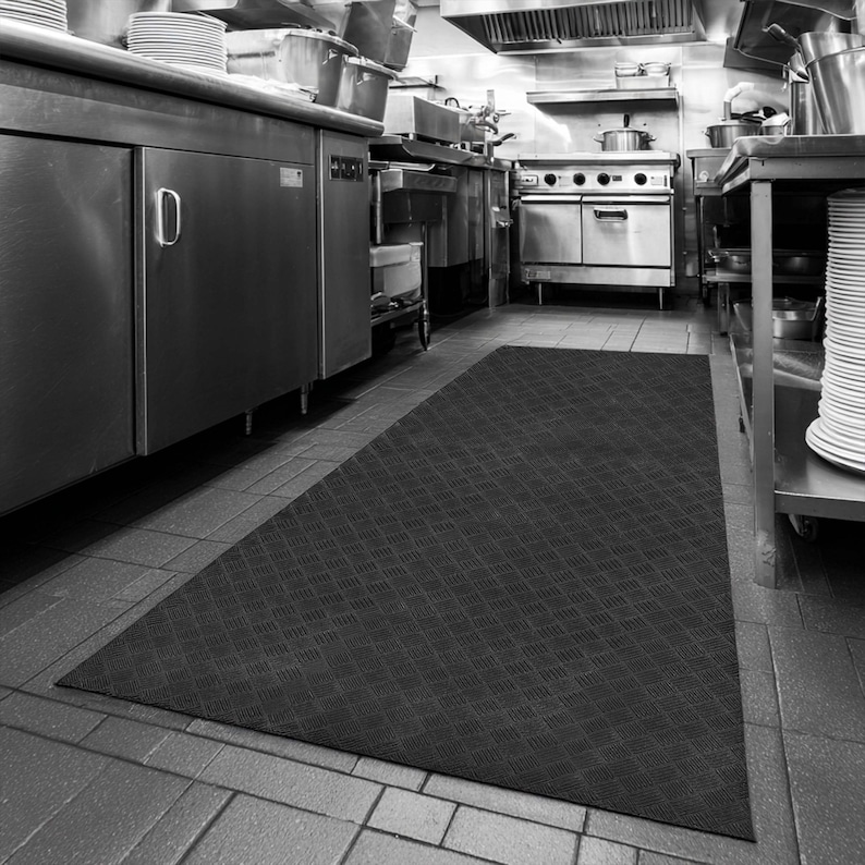 May include: Black rectangular anti-fatigue mat with a diamond pattern in a commercial kitchen. Stainless steel appliances and stacks of white plates are visible in the background. The mat is placed on a tiled floor.
