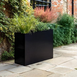 May include: A large, rectangular black planter box filled with various green plants and grasses. The planter is made of a solid material and sits on a stone patio. The background features a brick wall and lush greenery.