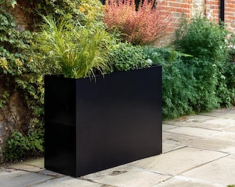 May include: A tall, rectangular black metal planter box with a smooth, matte finish. The planter is filled with various green grasses and leafy plants, creating a dense arrangement of foliage that extends above the top edge of the container.