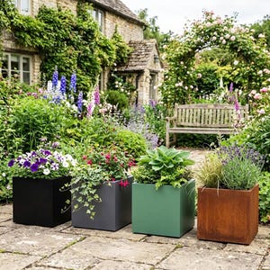 May include: Four square planters in black, grey, green, and rust-coloured metal arranged in a row on a stone patio. Each planter contains different plants, including purple and white petunias, red fuchsia, green hostas, and lavender with ornamental grass.
