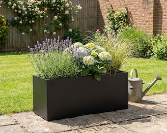 May include: A rectangular black metal planter box filled with purple lavender, white hydrangea flowers, and tall ornamental grass. A galvanized metal watering can sits on the stone patio next to the planter, which is positioned outdoors.