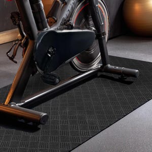 May include: Black exercise mat with a textured, basketweave pattern. A stationary exercise bike is partially visible, with a black frame and a red and black wheel. The mat is on a grey concrete floor.