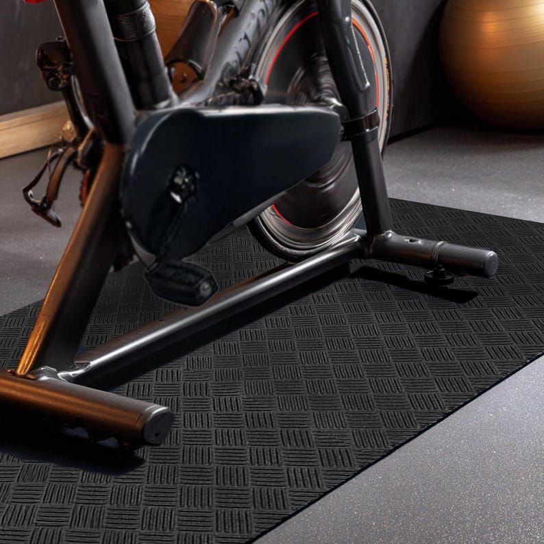 May include: Black exercise mat with a textured, basketweave pattern. A stationary exercise bike is partially visible, with a black frame and a red and black wheel. The mat is on a grey concrete floor.