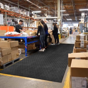 May include: A large black rubber mat with a diamond pattern is laid out in a warehouse. The mat is positioned in front of a blue table where several people are working. Cardboard boxes and pallets are visible.