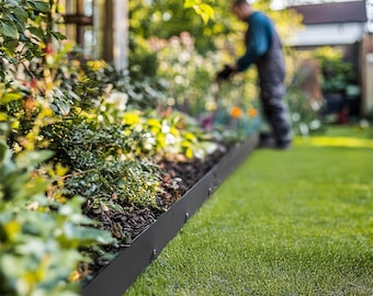 May include: A black metal garden edging borders a flower bed filled with green plants and dark soil. The edging is installed along a lush green lawn. A person is working in the background.