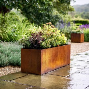 May include: A rectangular, weathered metal planter box filled with green and burgundy plants. The box has a rusty brown colour and sits on a stone patio. A second planter box is visible in the background, along with lush greenery.