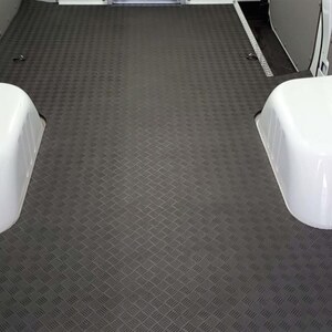 May include: Dark grey diamond-plate pattern flooring inside a vehicle. The flooring has a textured surface and is surrounded by white wheel arches. The floor is designed for durability and traction.