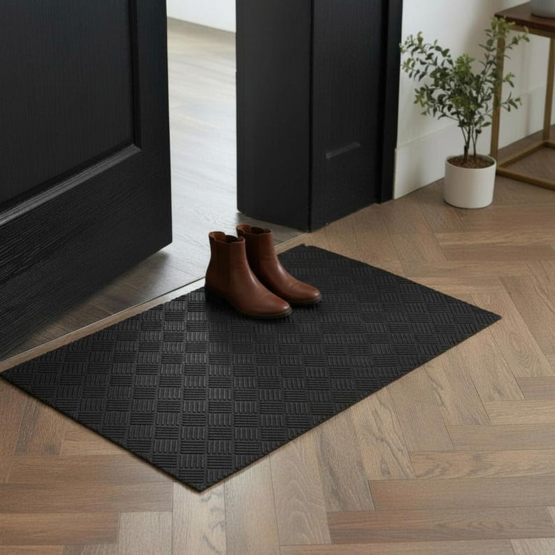 May include: A black rectangular doormat with a geometric pattern on a wooden floor. Two brown ankle boots are placed on the mat near a black door. The door frame is also black.