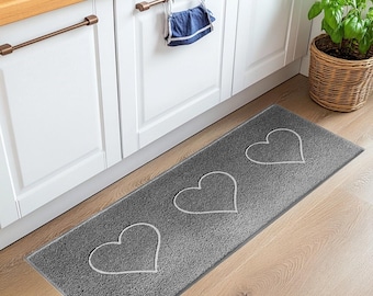 May include: A rectangular gray kitchen runner rug featuring three white outlined heart shapes centered along its length. The rug has a textured, durable surface and is positioned on a light wood floor in front of white kitchen cabinets.
